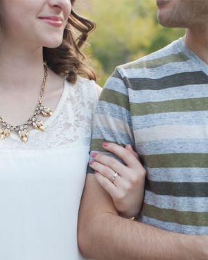 Couple posing outdoors, woman in white lace dress with statement necklace and engagement ring.