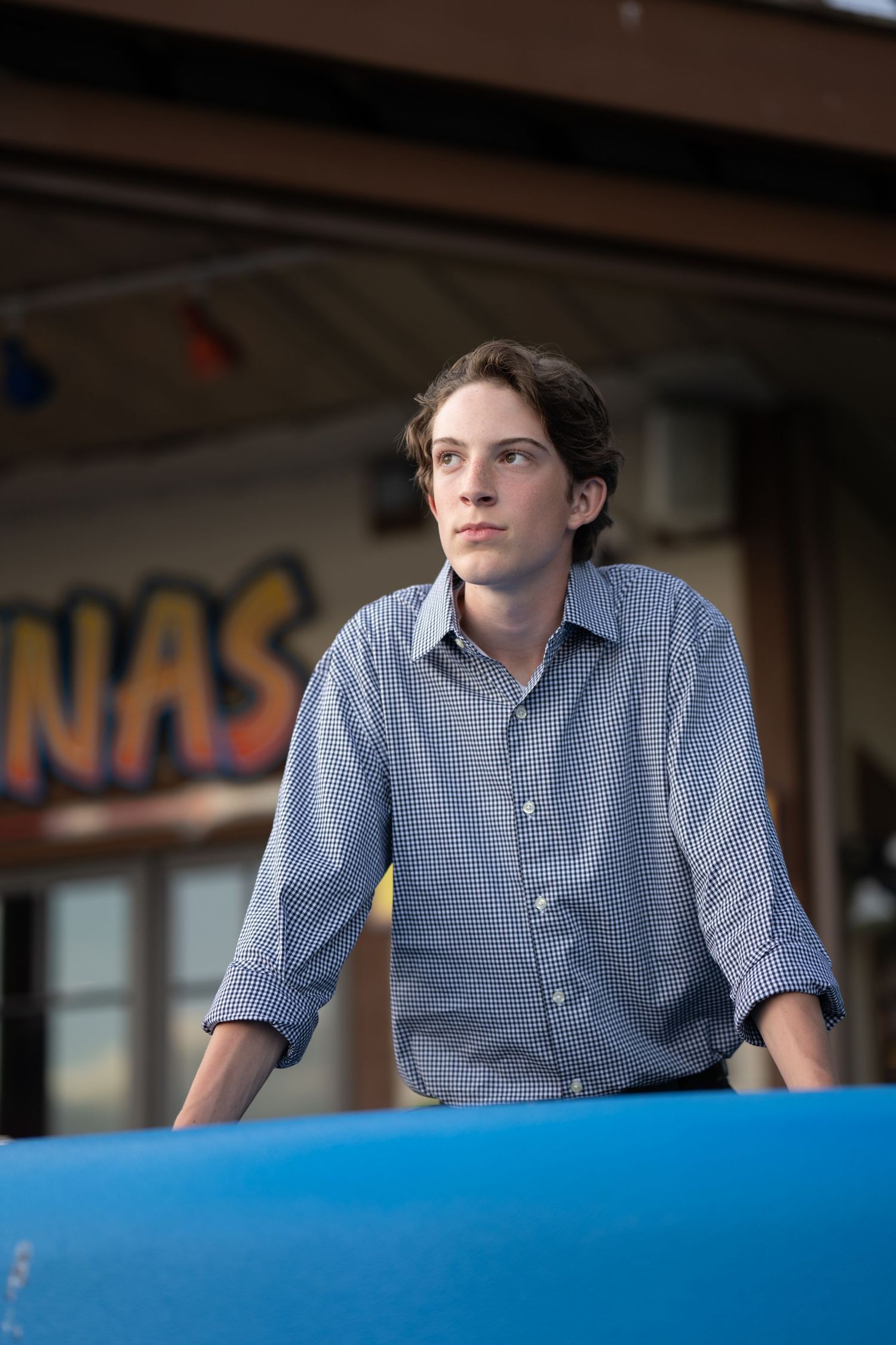 Person in blue checkered shirt leans on bright blue surface while looking off to the side.