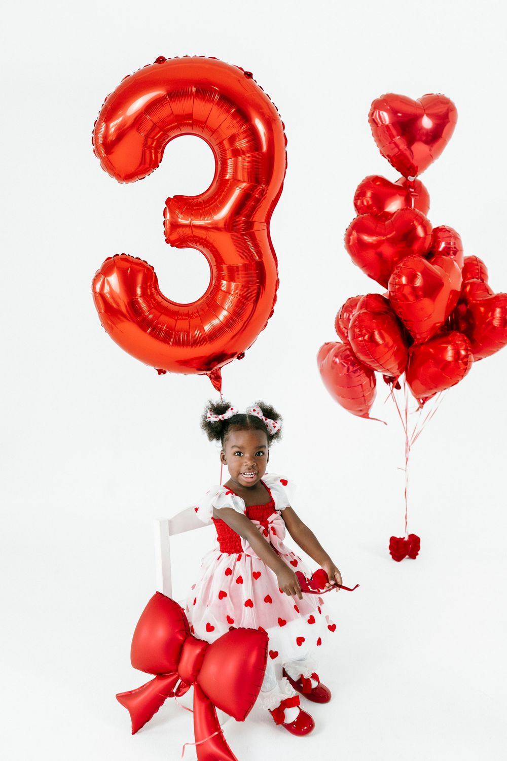 Child in white dress poses with red heart balloons and number 3 balloon for birthday celebration photoshoot.