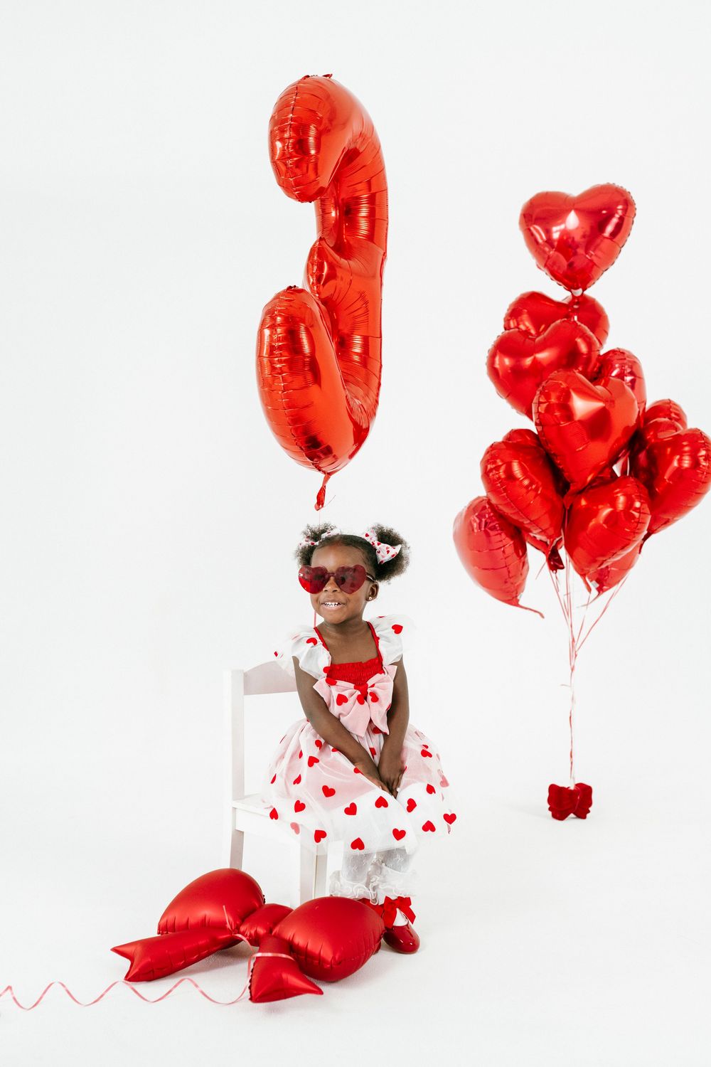 A child in a white dress with red hearts poses with red heart-shaped balloons against a white background.