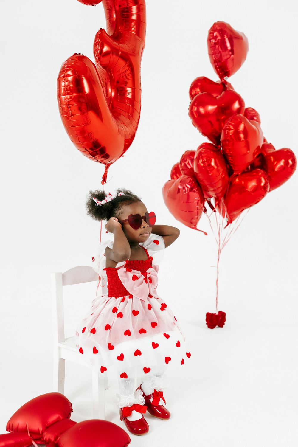 A child in a white and red heart-patterned dress poses with red heart-shaped balloons against a white backdrop.