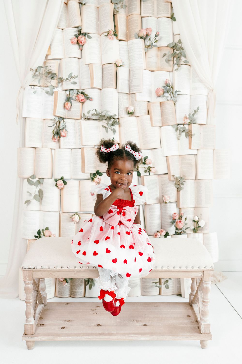 Three photos of a young child in a white dress with red hearts sitting on a rustic white bench with floral decorations.
