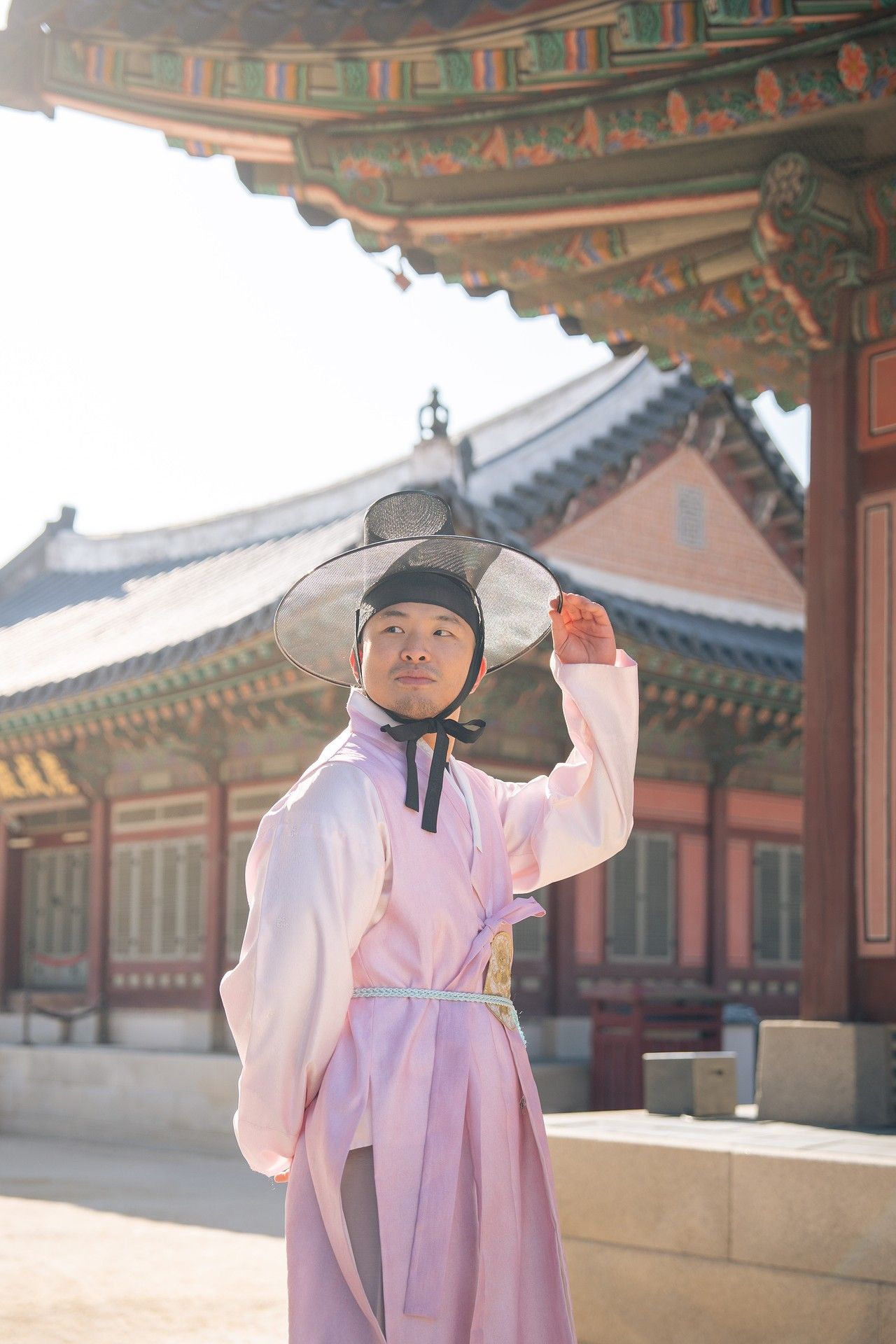 A person in traditional Korean hanbok dress stands beneath ornate temple architecture on a sunny day.