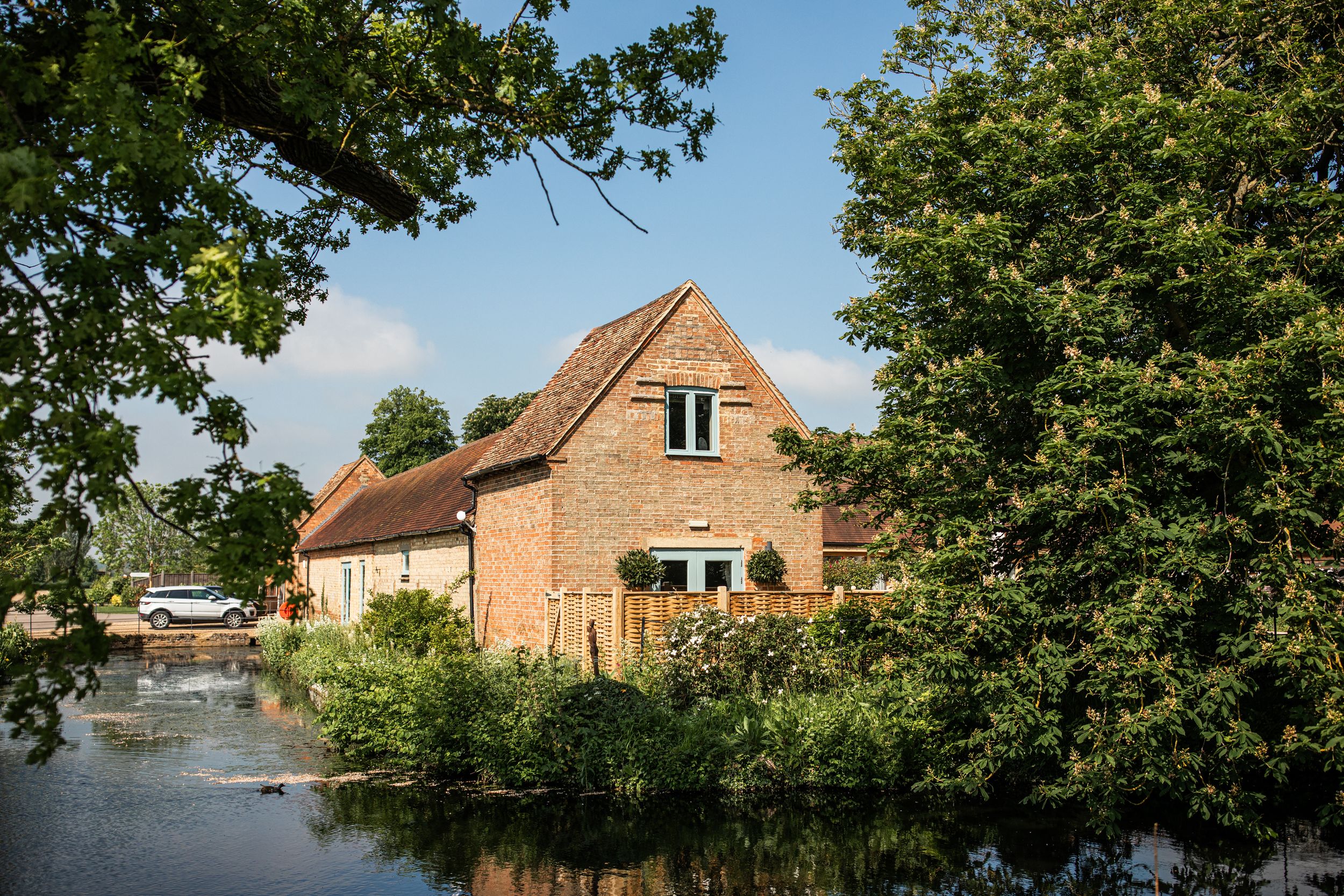 Charming rustic brick wedding venue beside a tranquil river surrounded by mature trees in summer.