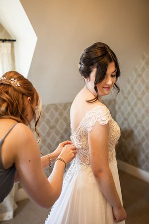 Bridesmaid carefully fastening buttons on back of bride's lace off-shoulder wedding gown indoors.