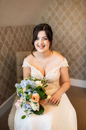 Radiant bride in lace off-shoulder gown holding pastel blue and peach floral bouquet while seated.