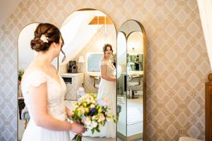 Bride in white lace gown holding floral bouquet reflected in elegant gold arch mirror during wedding preparations.