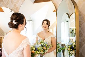 Beautiful bride in lace wedding dress admiring her reflection in tall gold arched mirror with bouquet.