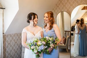 Smiling bride and bridesmaid in blue dress holding wildflower bouquets together in bright wedding preparation room.