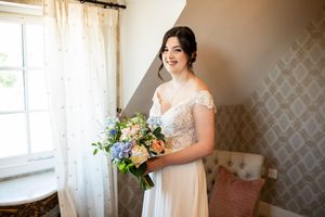 Beautiful bride in off-shoulder lace wedding gown holding pastel floral bouquet standing by window in natural light.