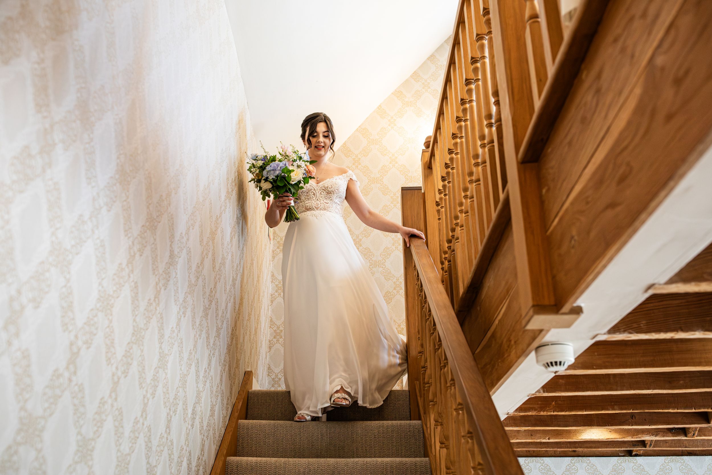 Elegant bride in flowing white gown descending wooden staircase holding colorful bouquet in wallpapered venue.