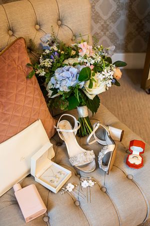 Bridal flat lay featuring blue hydrangea bouquet, jeweled heels, jewelry, and ring box on chair.