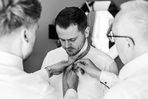 Black and white photo of groom being helped with his tie by two men before the wedding ceremony.