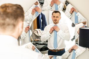 Groom adjusting his blue tie in a geometric mirror reflection while getting ready for his wedding day.