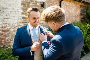 Smiling groom in blue suit receives boutonniere pinned to lapel by groomsman outside brick wall venue.
