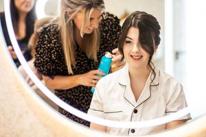 Hairstylist spraying finishing touches on brunette bride's elegant updo reflected in lit vanity mirror.