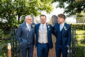 Three men in coordinating navy blue wedding suits smiling together in a sunlit garden setting.