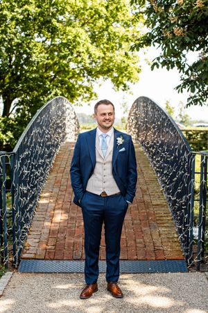 Handsome groom in navy suit and beige waistcoat poses confidently on brick bridge surrounded by trees.