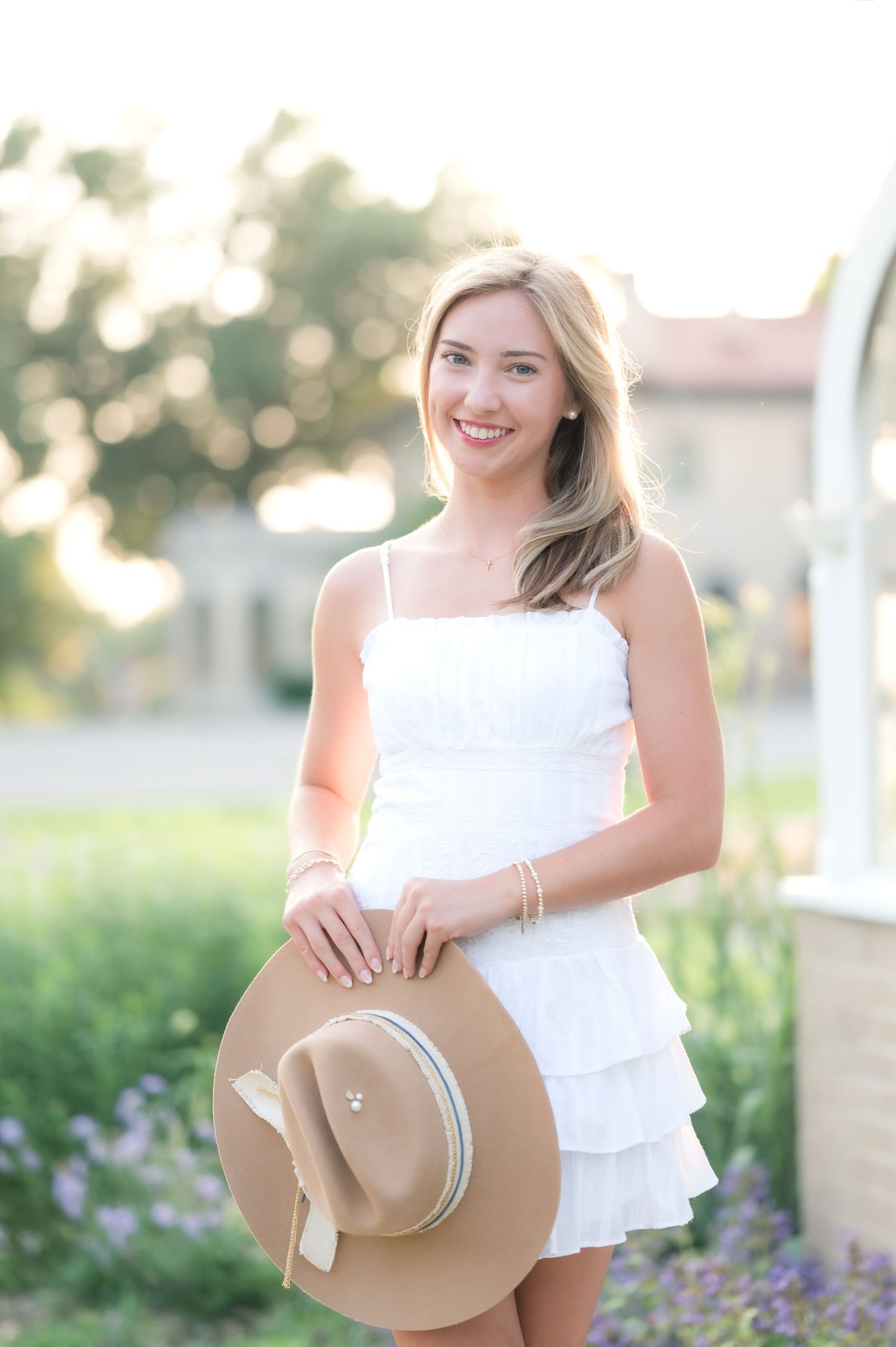 A high school senior in a white ruffle dress and sun hat poses playfully in a garden with purple flowers during golden hour.