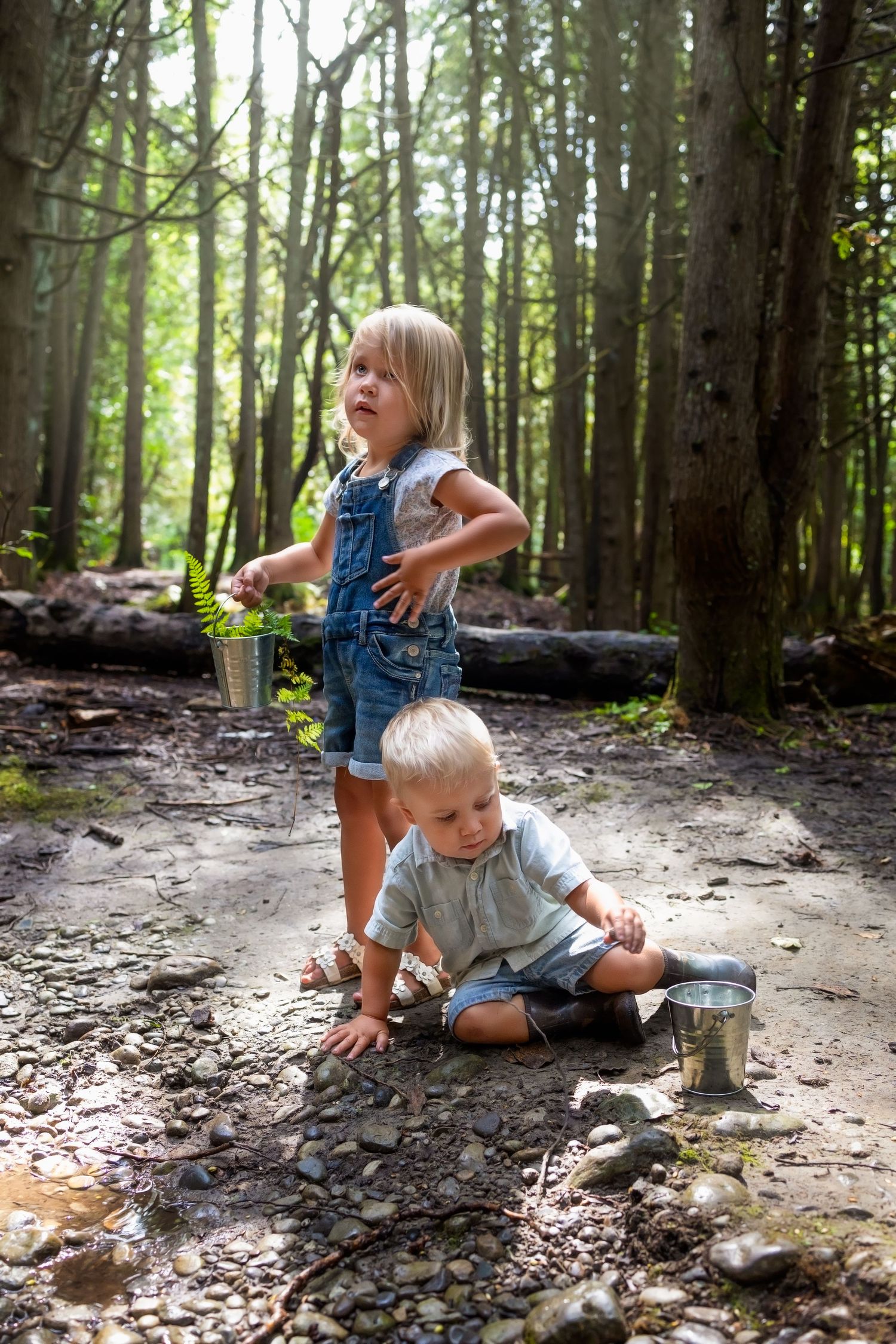 Two children play and explore together on a dirt path in a sunlit forest while wearing casual summer clothes.