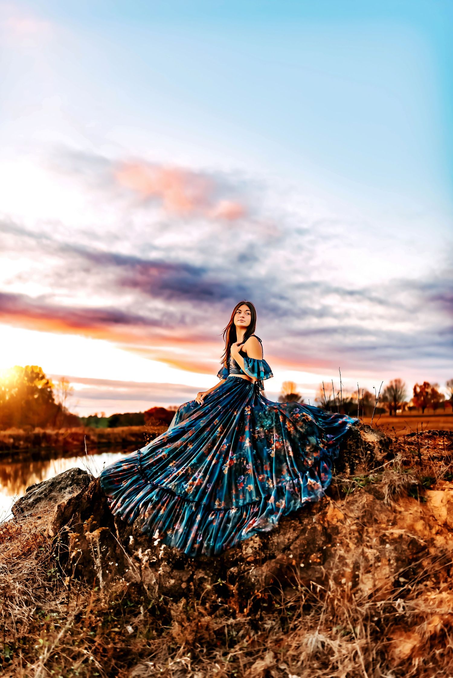 A person in a flowing rust-colored dress poses gracefully on a grassy hillside at sunset with wooden fencing in the background.