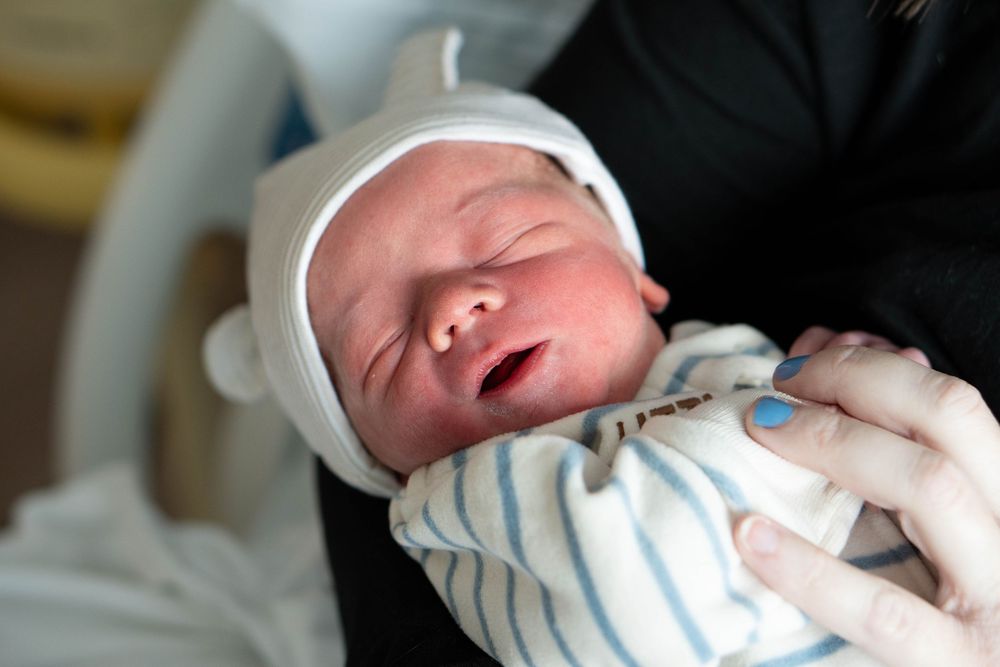 A newborn baby with mouth open sleeps peacefully while being held in a hospital setting.
