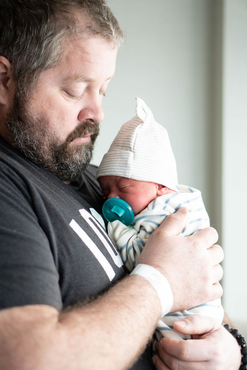 Bearded father tenderly holding his newborn baby with a pacifier and striped knit hat in a hospital room.