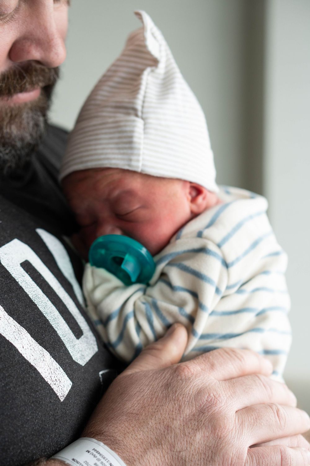 Close-up of a newborn baby with a teal pacifier sleeping against a bearded fathers chest in hospital.