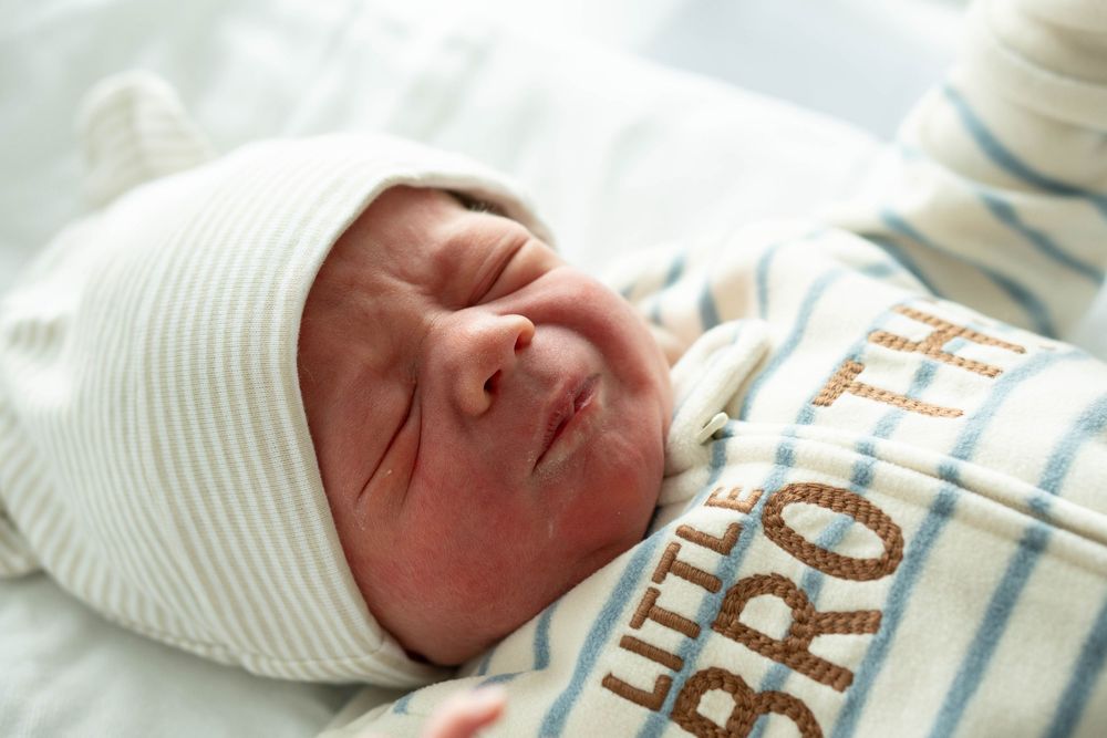 Peaceful newborn baby wearing a white hat and Little Brother striped onesie resting in hospital bassinet.