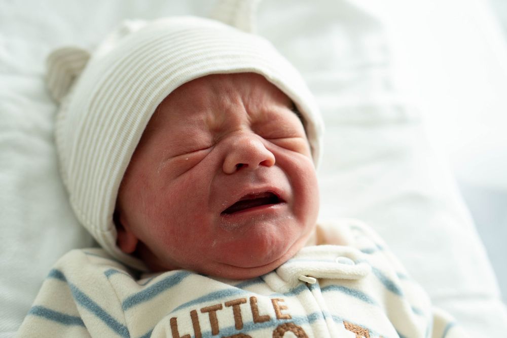 Crying newborn baby in white bear ear hat and Little Brother onesie in hospital bassinet.