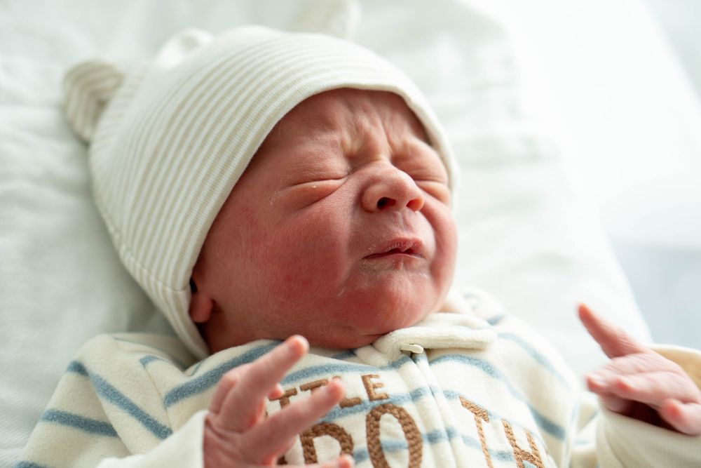 Fussy newborn baby with raised hands wearing white bear hat and Little Brother onesie in hospital.