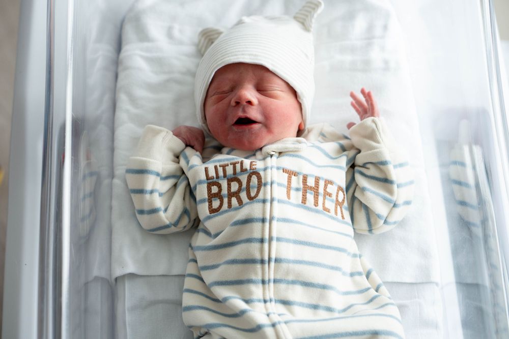 Wide-eyed newborn baby in white bear ear hat and Little Brother onesie lying in clear hospital bassinet.