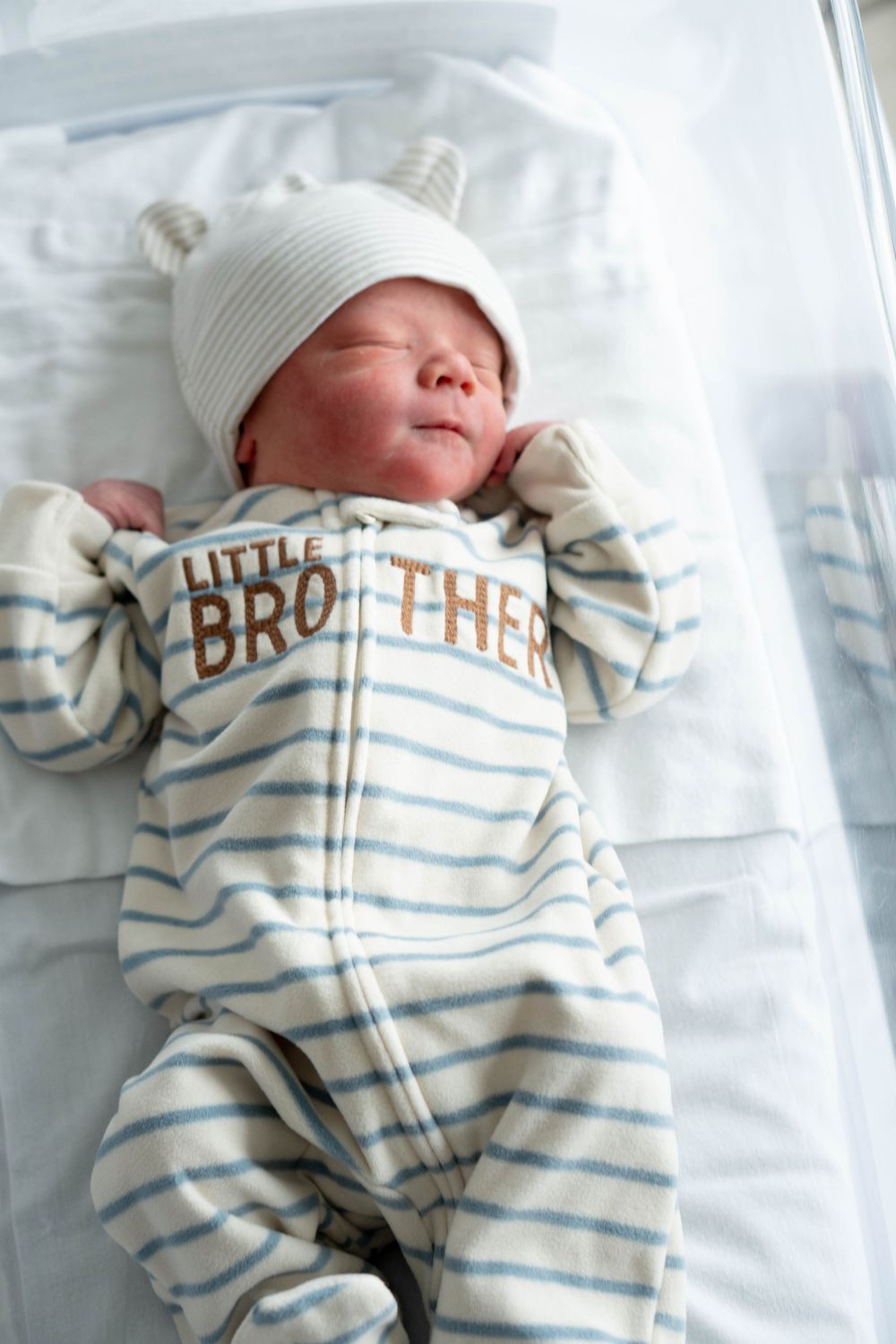 Full-body view of sleeping newborn in Little Brother striped onesie and white bear hat in hospital bassinet.