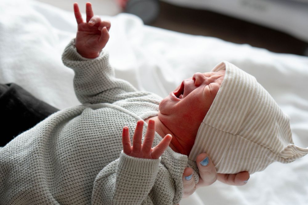 Crying newborn baby in gray onesie and striped hat raising tiny hands while lying on white bedding.