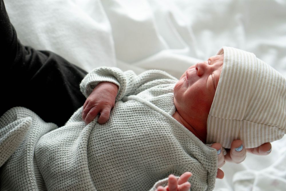 Newborn baby wrapped in gray knit blanket wearing striped hat resting in adult arms on white bedding.