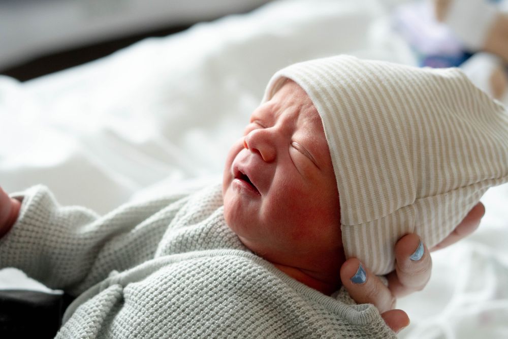 Close-up of crying newborn in gray onesie and striped hat being gently held by hand with blue nails.