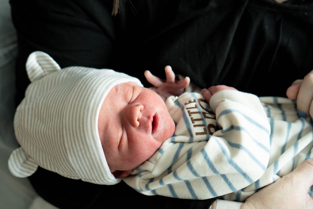 A sleeping newborn baby in a striped outfit and bear ear hat is cradled in someone's arms.
