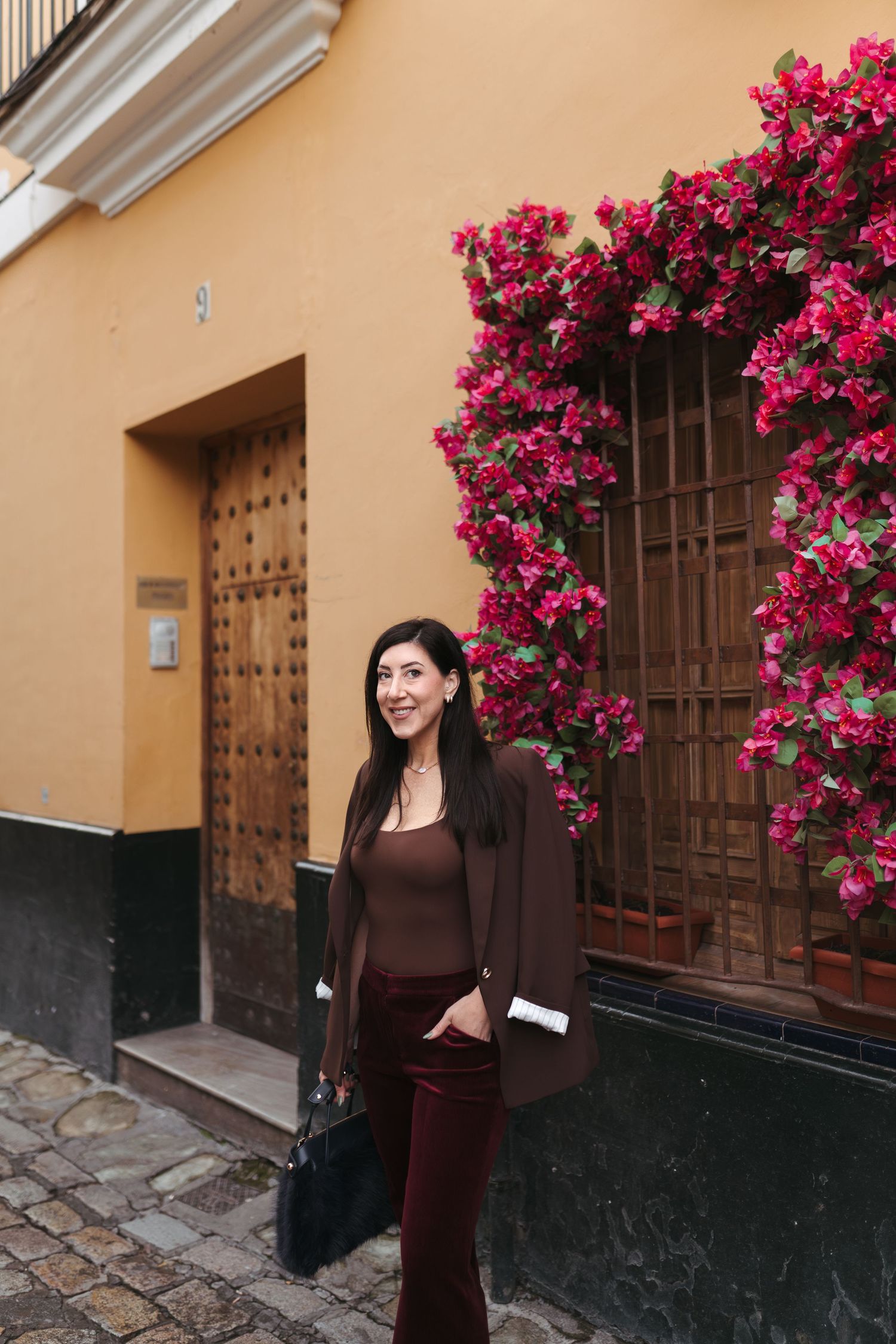 Woman in brown outfit poses by vibrant pink bougainvillea flowers against yellow building wall.