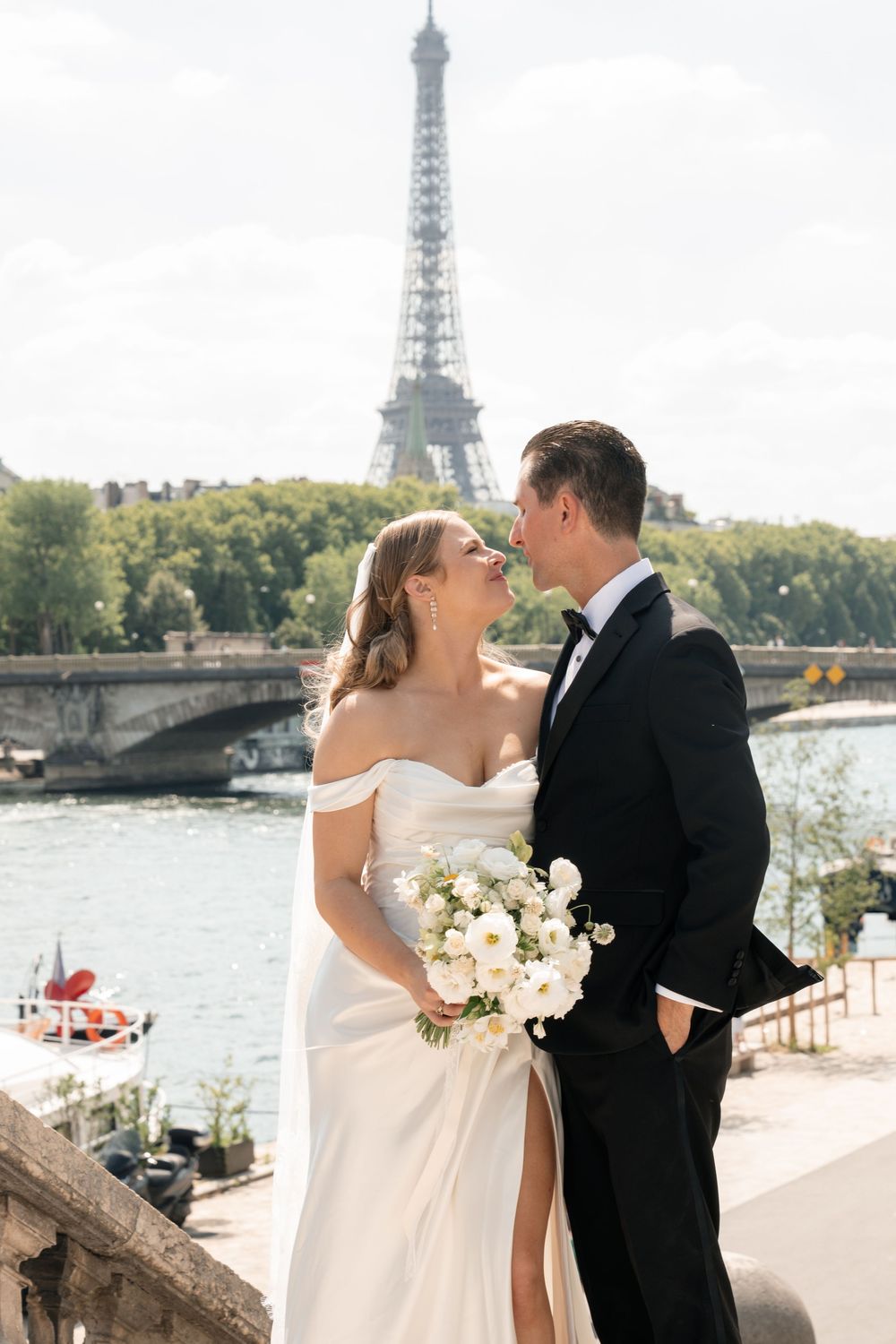 Newlyweds share intimate moment by Seine River with Eiffel Tower in background.