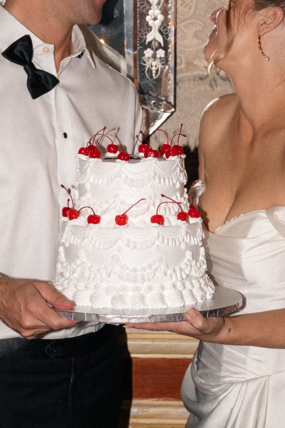 A wedding couple cutting into their white tiered cake decorated with red cherries around the edge.