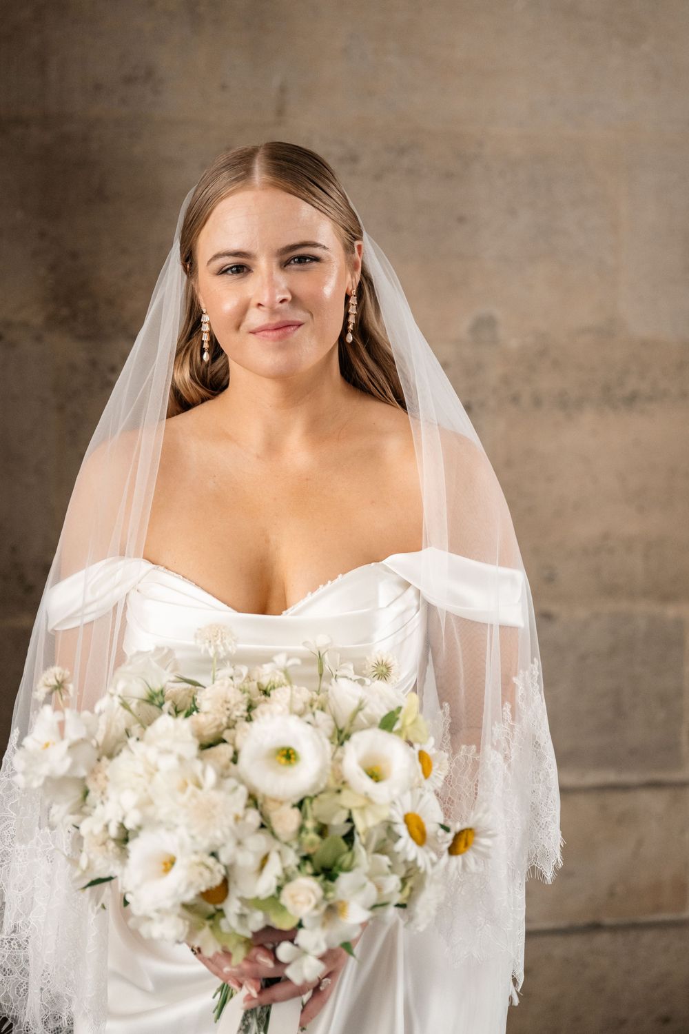A bride in white dress holds elegant white bouquet against textured wall.
