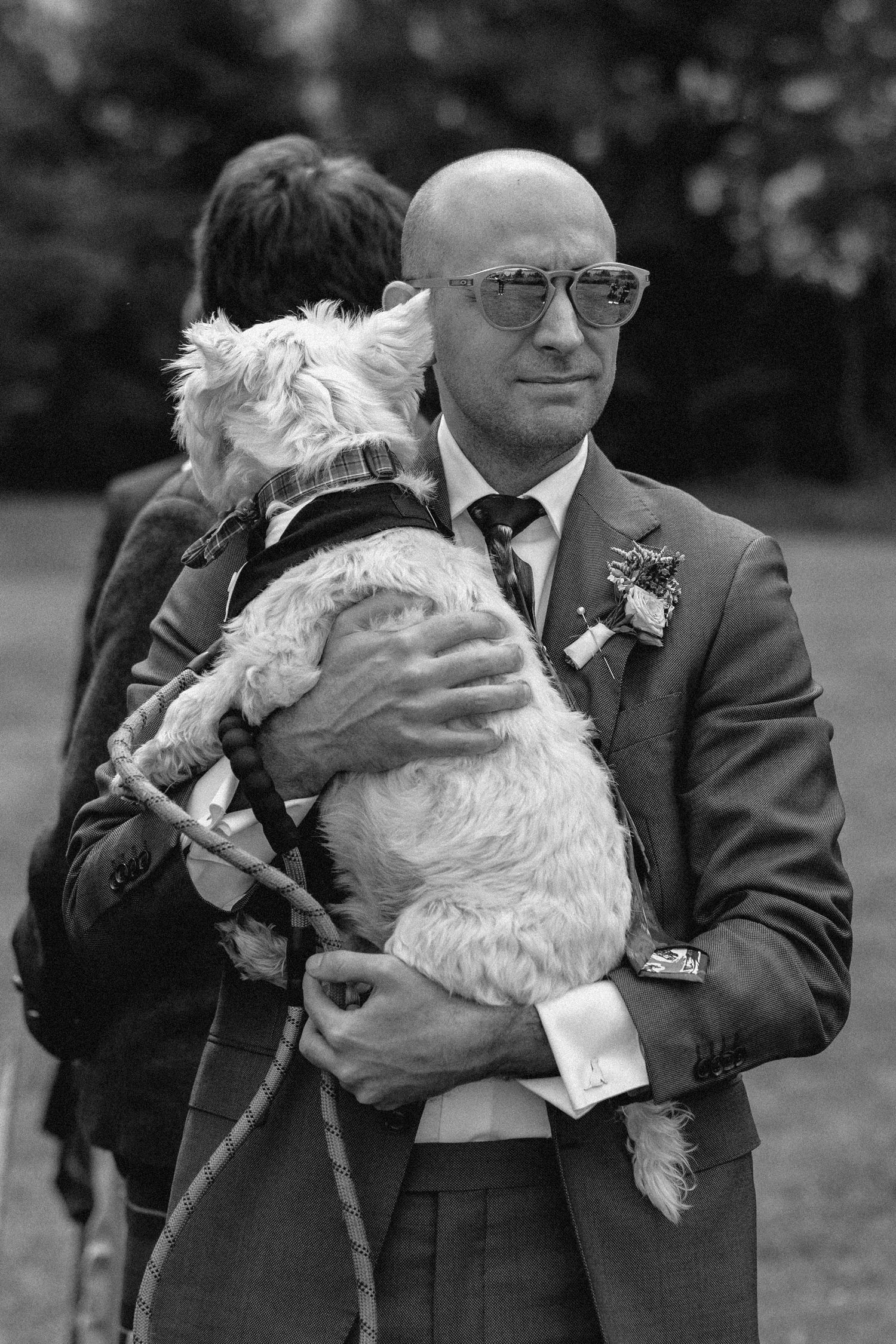 Black and white photo of a person in a suit and glasses holding a fluffy white dog at a formal outdoor event.