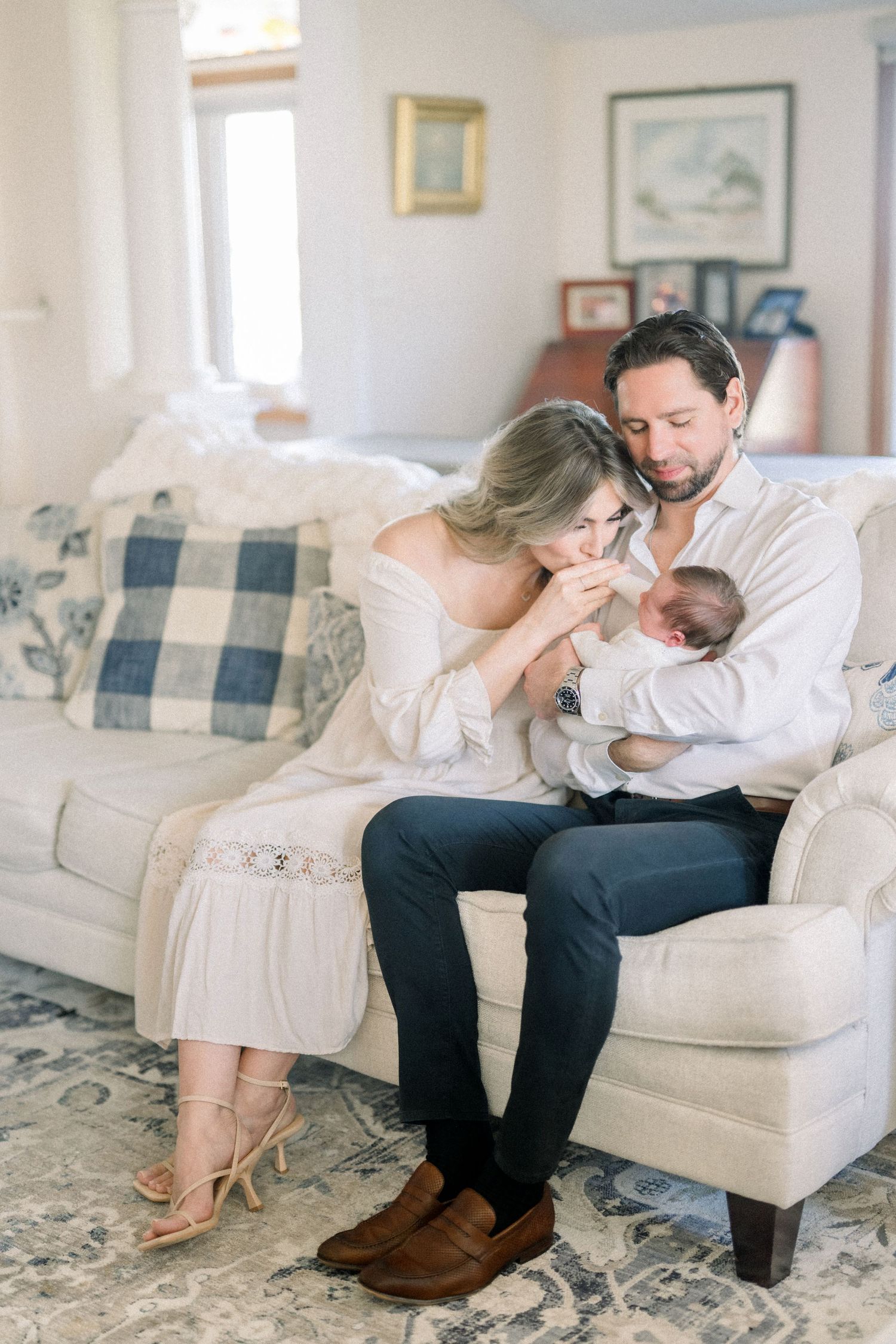 A couple sits together on a cream-colored couch with their newborn baby in a bright, cozy living room.