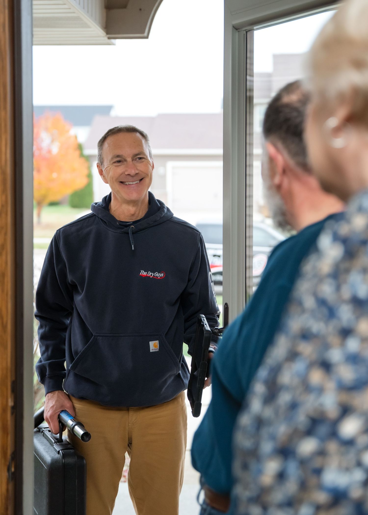A person wearing a navy blue Carhartt hoodie and khaki pants stands smiling in a doorway during autumn.