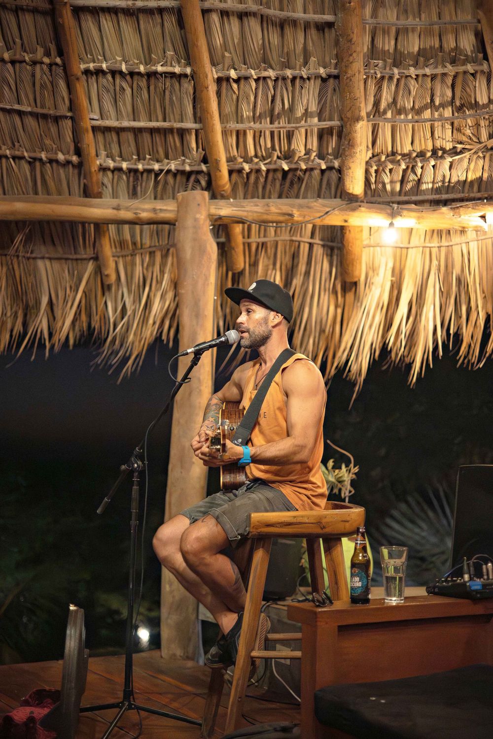 People enjoying drinks at a rustic tiki bar with thatched palm roof during evening hours.