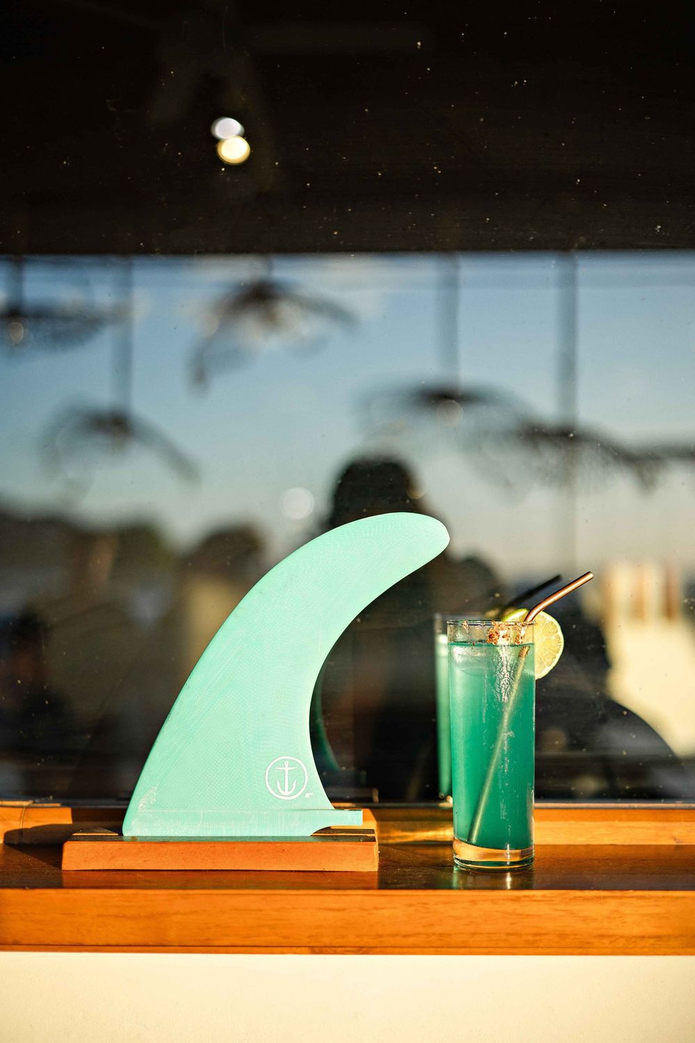 A light green surfboard fin sits on a wooden table with a blue cocktail glass in the background at sunset.