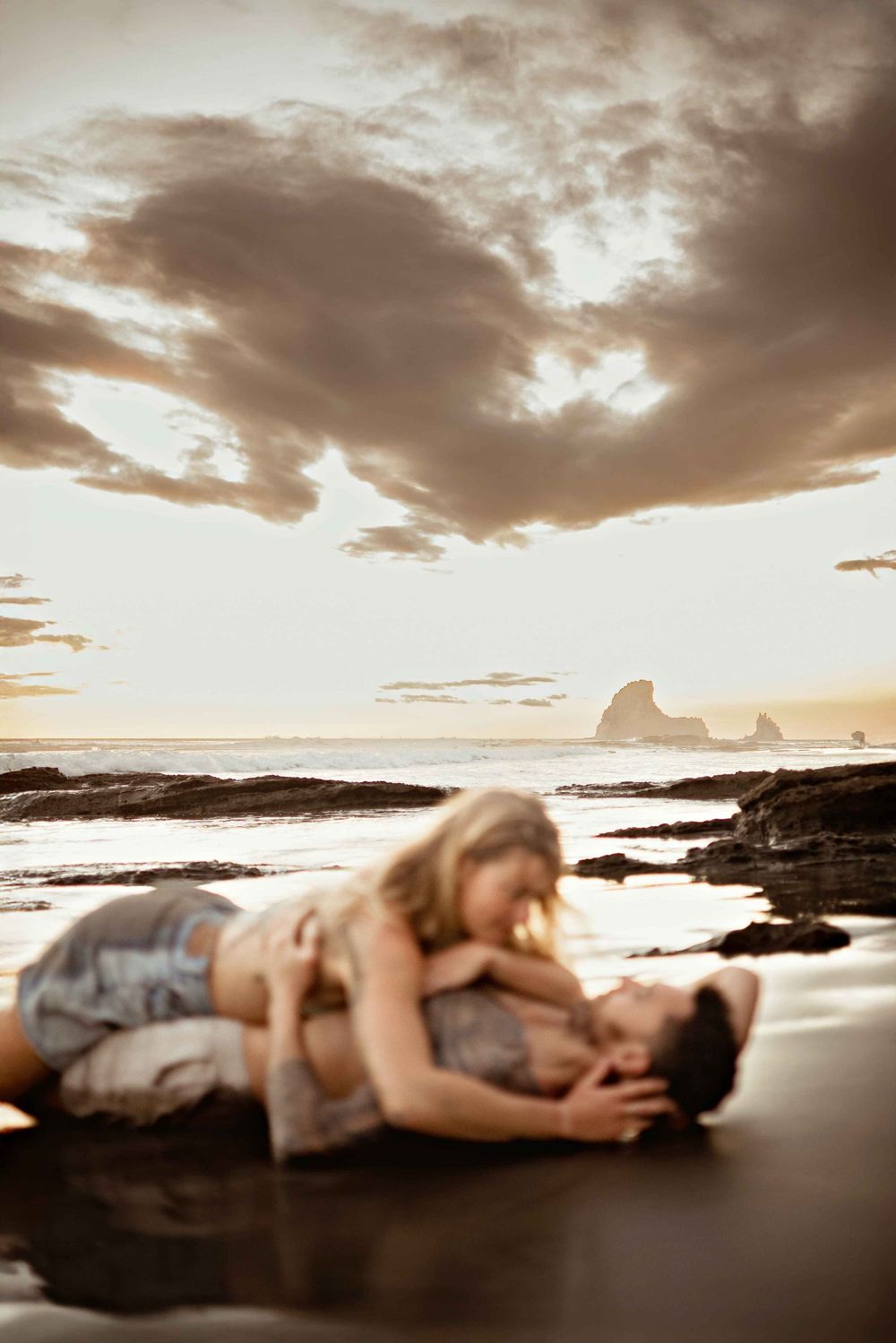A figure rests peacefully on dark coastal rocks at sunset with dramatic cloudy skies and ocean waves in the background.