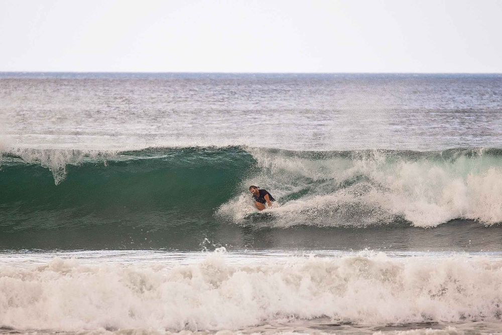 A surfer ducks under a breaking ocean wave with white spray against a misty gray horizon.