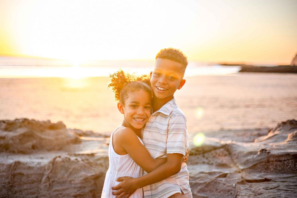 Couple embraces on rocky beach at sunset while ocean waves crash behind them creating a romantic silhouette.