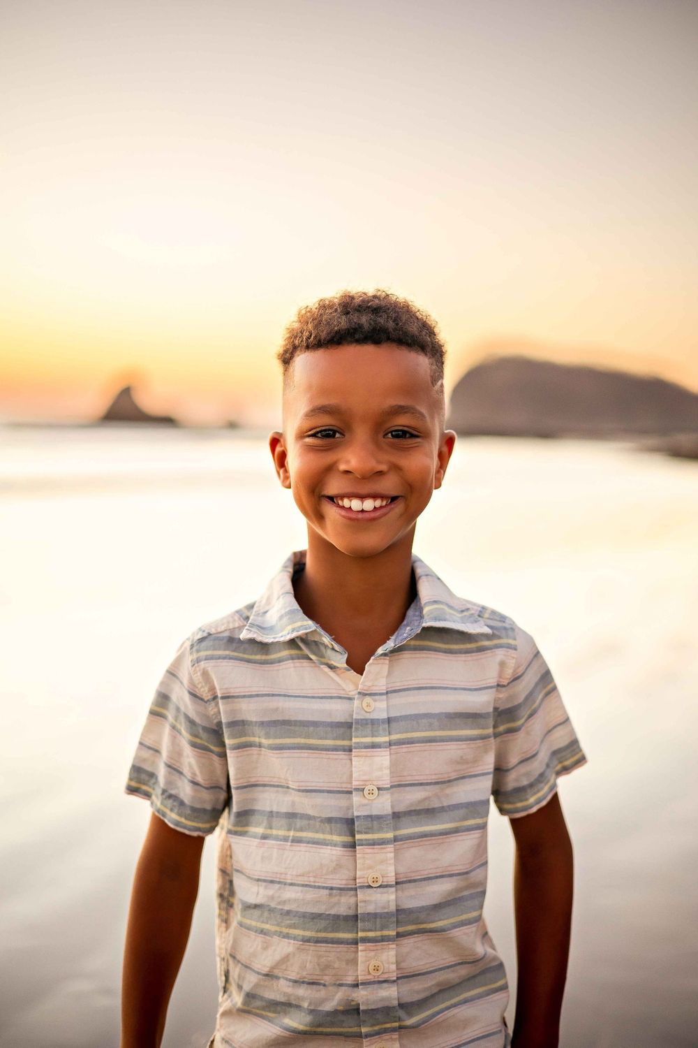 A young person smiles at the beach during golden hour with mountains visible in the distance.